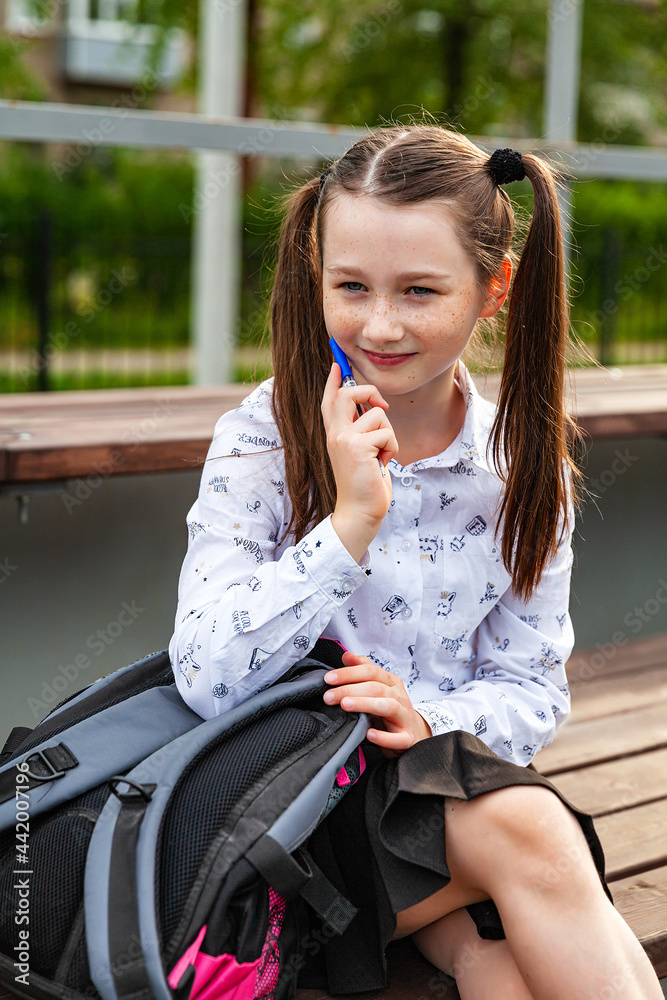 A smiling schoolgirl in a white blouse and black skirt, sitting on a bench on the school sports field, holding a pen. Portfolio, education, thought, dreams A smiling schoolgirl in a white blouse and black skirt, sitting on a bench on the school sports field, holding a pen. Portfolio, education, thought, dreams