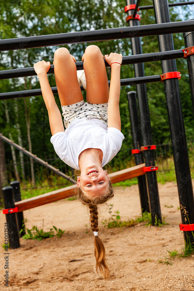 Obraz premium A girl in sports uniform hangs upside down on the turnstile. Sports ground, charging, warm-up, daytime, open air
