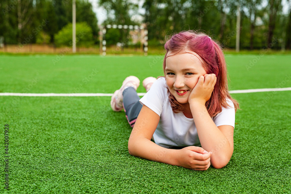 A happy girl lies on the grass on the playground. Daytime, stadium, open air