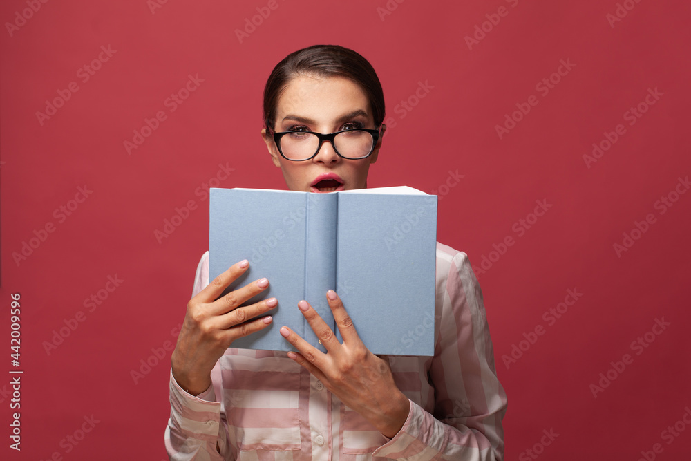 Portrait of a pretty young surprised woman hiding behind an open book and looking away on red background Portrait of a pretty young surprised woman hiding behind an open book and looking away on red background