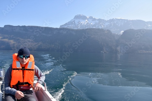 Boy on a motorboat on lake Lucerne in Switzerland. He is wearing orange life jacket and sun glasses. On the background there are snow-capped Alp mountains.