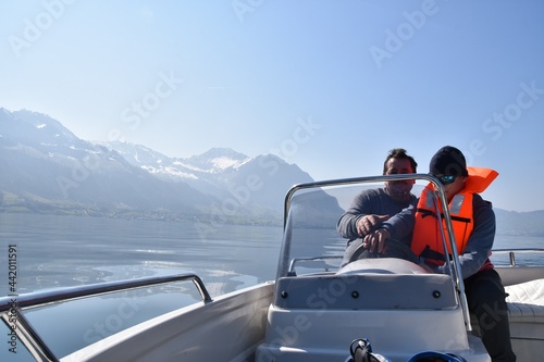 Father teaching his son how to drive a motorboat on lake Lucerne. The son is wearing orange life jacket. There is a lot of copy space on the background.