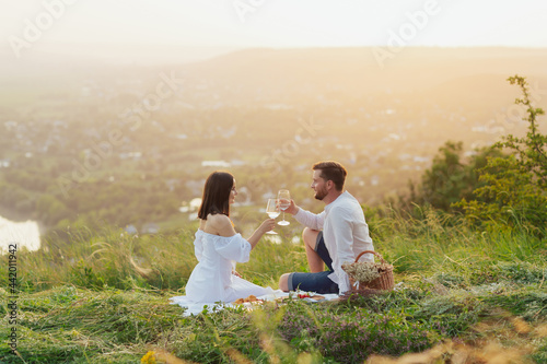 Couple in love sitting on a picnic plaid on the hill and drinking wine and enjoying the sunset.