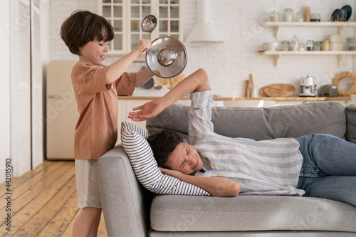 Foto Naughty kid rumbling kitchenware while tired mother try to sleep on sofa in living room