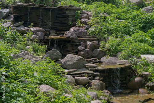 A three-level waterfall with slowly flowing, falling down and bubbling water flowing over stones and small stone slabs among boulders, green grass and fluffy bushes.