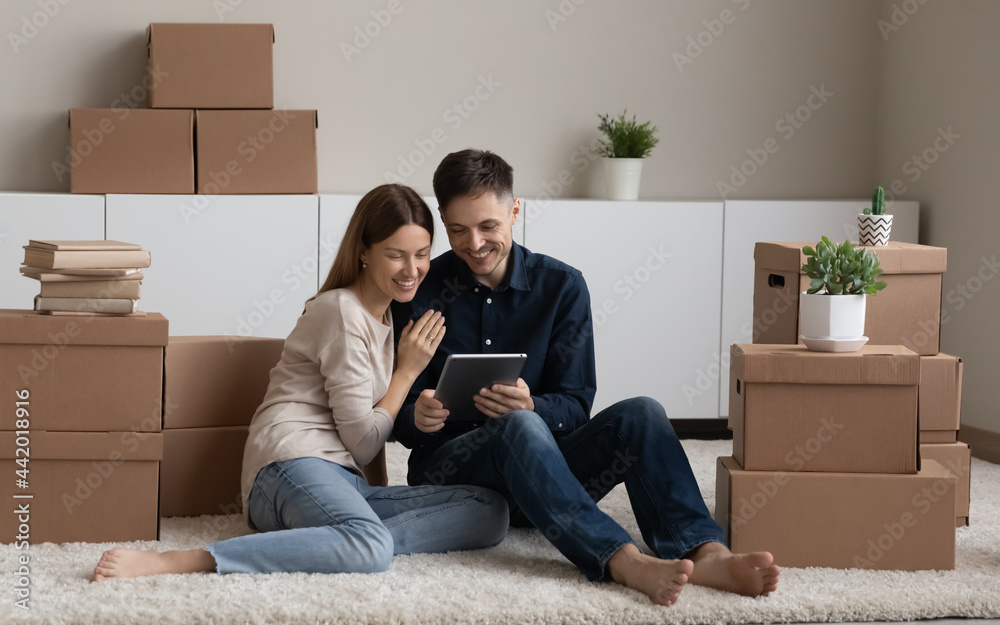 Smiling young Caucasian man and woman renters sit on floor in new home near boxed packages use tablet gadget. Happy couple tenants hold pad device browse internet search apartment for rent online. Smiling young Caucasian man and woman renters sit on floor in new home near boxed packages use tablet gadget. Happy couple tenants hold pad device browse internet search apartment for rent online.