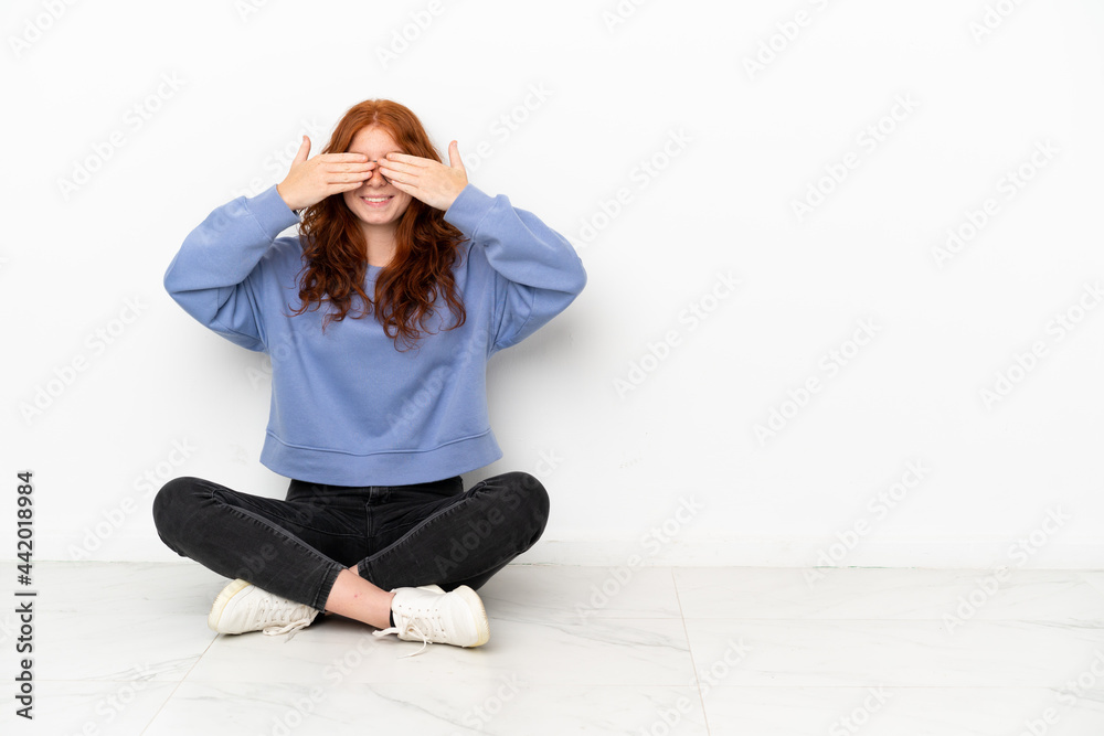 Teenager redhead girl sitting on the floor isolated on white background covering eyes by hands Teenager redhead girl sitting on the floor isolated on white background covering eyes by hands