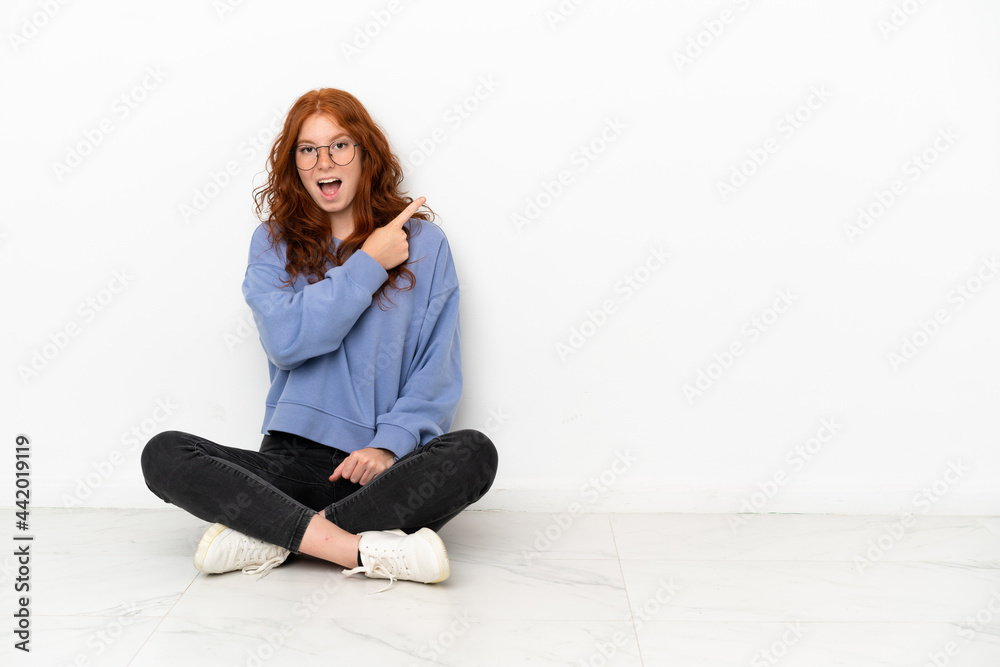 Teenager redhead girl sitting on the floor isolated on white background surprised and pointing side Teenager redhead girl sitting on the floor isolated on white background surprised and pointing side