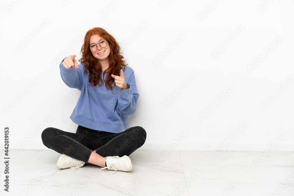 Teenager redhead girl sitting on the floor isolated on white background points finger at you while smiling Teenager redhead girl sitting on the floor isolated on white background points finger at you while smiling