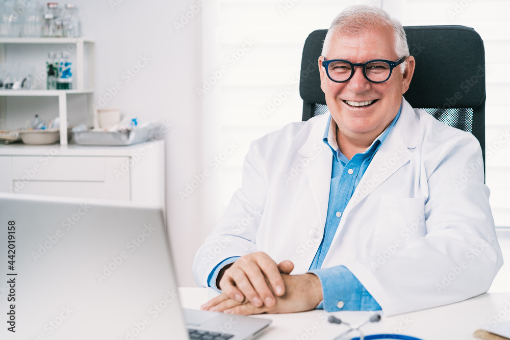 Positive senior physician working on laptop in hospital