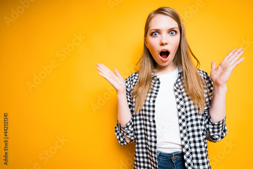 A cute girl in a plaid shirt is surprised and spreads her hands while opening her mouth