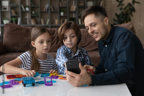 Happy young Caucasian father play toys with two teen kids use modern smartphone have webcam call on device. Smiling loving dad and children involved in funny game with blocks at home together.