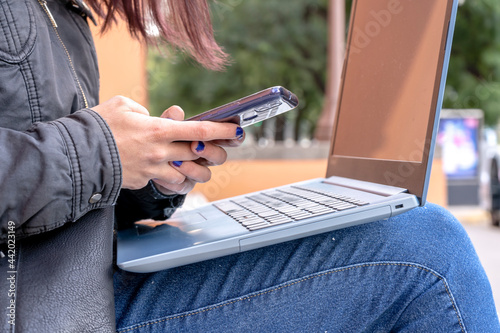 Young Latin woman answering a message with the cell phone while working with the laptop sitting on a staircase. Concept of comunications work, technology, IT. photo