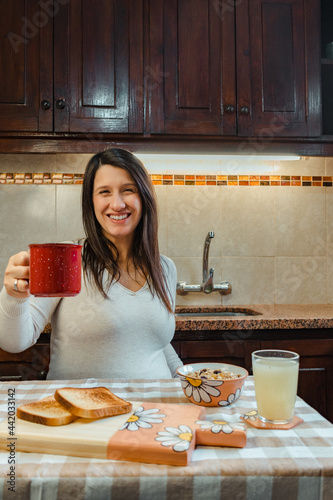 Vertical shot of a Hispanic woman raising a cup of coffee as a sign of toast while looking at the camera and smiling during a breakfast at home