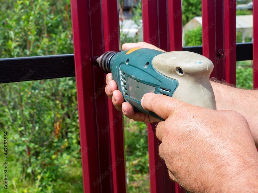 Construction of a fence in the garden. A man using a drill screws a roofing screw into a metal picket fence. The concept of building and upgrading a private territory. Construction of a fence in the garden. A man using a drill screws a roofing screw into a metal picket fence. The concept of building and upgrading a private territory.