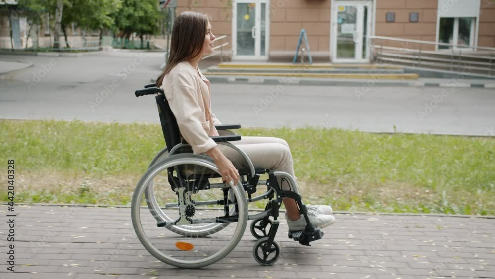 Side view of beautiful disabled woman moving in wheelchair along city street smiling enjoying ride and fresh air. Handicapped people and mobility concept. Side view of beautiful disabled woman moving in wheelchair along city street smiling enjoying ride and fresh air. Handicapped people and mobility concept.