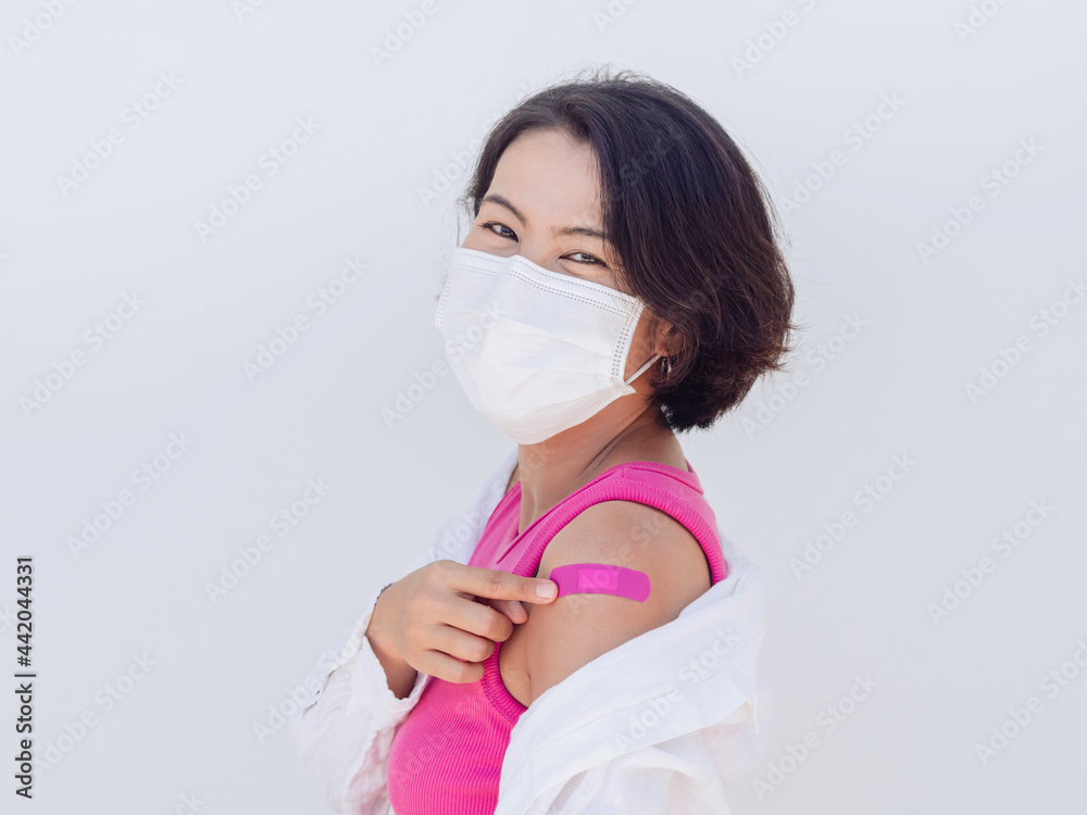 Vaccinations, vaccinated people concept. Happy Asian women wearing face mask and pink sleeveless shirt pointing bandage on own shoulder with smile after vaccination treatment on white background.