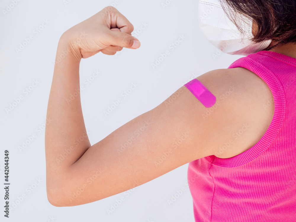 Vaccinations, bandage on vaccinated people concept. Pink bandage on female shoulder who wearing pink sleeveless and face mask and showing strong gesture with fist and arm after vaccination treatment. Vaccinations, bandage on vaccinated people concept. Pink bandage on female shoulder who wearing pink sleeveless and face mask and showing strong gesture with fist and arm after vaccination treatment.