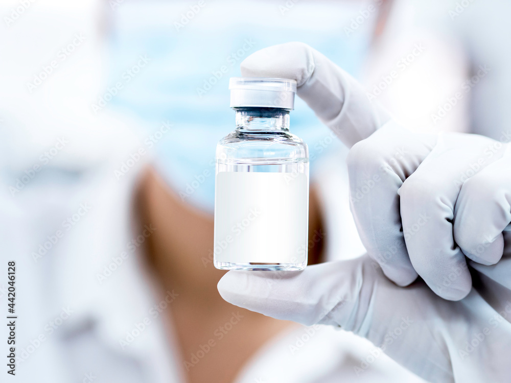 Medical vaccine vial with white blank label in scientist hand wearing face mask and white rubber gloves with copy space. Close up image of hand holding vaccine glass bottle for injection. Medical vaccine vial with white blank label in scientist hand wearing face mask and white rubber gloves with copy space. Close up image of hand holding vaccine glass bottle for injection.