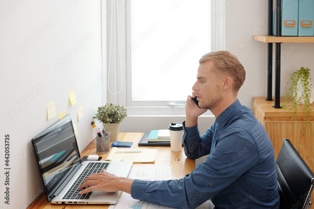 Pensive young entrepreneur talking on phone with colleagues when filling form in laptop at his desk Pensive young entrepreneur talking on phone with colleagues when filling form in laptop at his desk