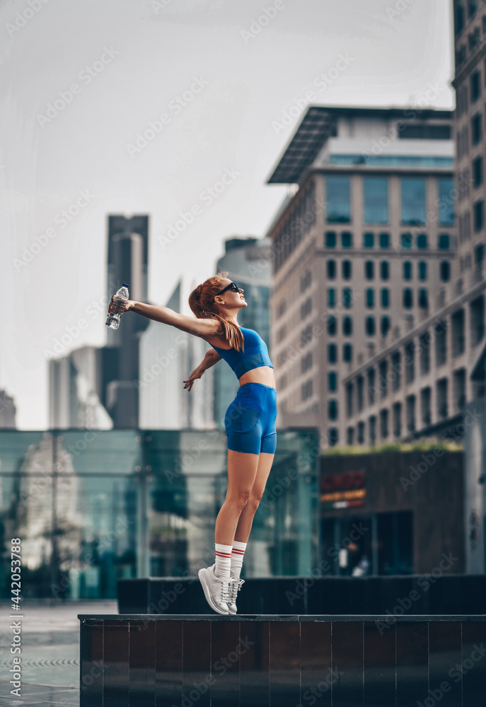 Fototapeta premium Happy slim, flexible, athletic red-haired woman in summer sportswear shorts and top stands sideways on tiptoe, holding hands up on city street in Dubai. Side view