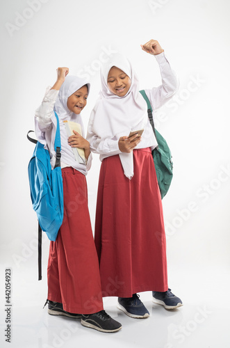 two veiled girls wearing elementary school uniforms using a mobile phone together with a backpack on an isolated background