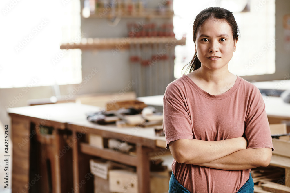 Portrait of young female carpenter crossing arms and looking at camera when standing in workshop Portrait of young female carpenter crossing arms and looking at camera when standing in workshop