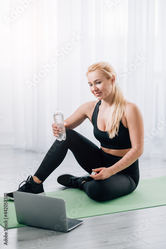 Refreshing body. Sportive woman. Online training. Home fitness. Happy slim lady in black sportswear sitting yoga mat holding bottle of clear water looking laptop in light room interior.