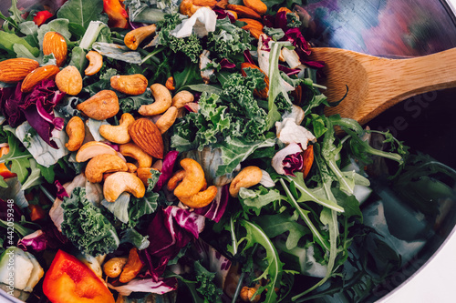 Salad with fresh vegetables, leaves, cashews, hazelnuts and almonds in a metal bowl is mixed with a wooden spoon