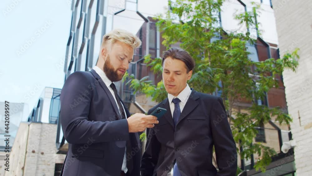 Confident businessman and his colleague in front of modern office building. Financial investors are using smartphone device and talking. Banking and business. Confident businessman and his colleague in front of modern office building. Financial investors are using smartphone device and talking. Banking and business.