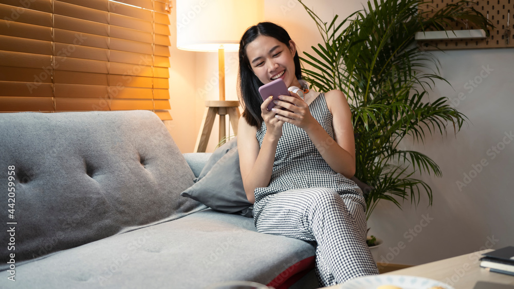 Obraz premium Smiling female sitting on sofa at home and using mobile phone.