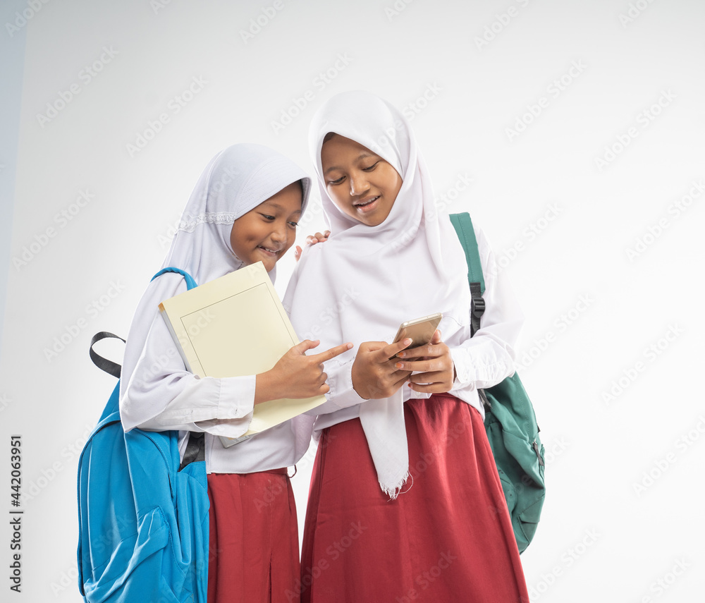 two veiled girls wearing elementary school uniforms while using a mobile phone together with a backpack and a book with an isolated background two veiled girls wearing elementary school uniforms while using a mobile phone together with a backpack and a book with an isolated background