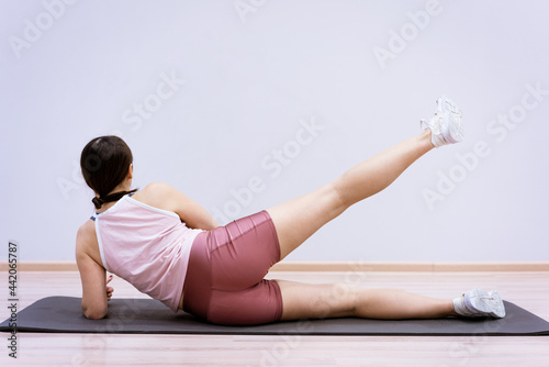 Fototapeta Naklejka Na Ścianę i Meble -  A woman of Caucasian ethnicity in sportswear is engaged in fitness at home against the background of a wall. Healthy lifestyle and slim body concept