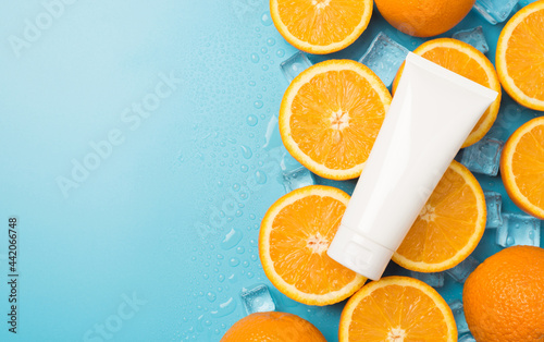 Top view photo of white tube without label on ice cubes water drops halves and whole oranges on isolated pastel blue background with blank space on the left
