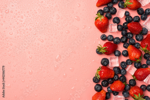 Top view photo of scattered strawberries with leaves blueberries and ice cubes on the right water drops on isolated light pink background with copyspace on the left