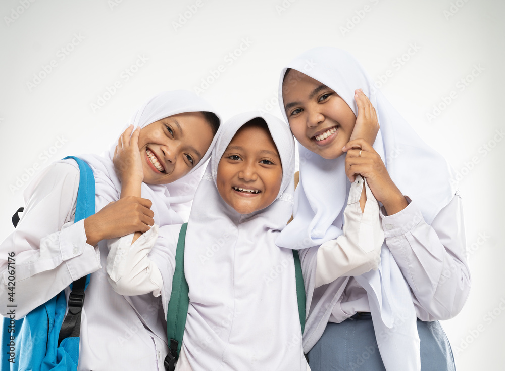 three girls in headscarves wearing school uniforms stand smiling with affectionate gestures of each other while carrying backpacks against an isolated background three girls in headscarves wearing school uniforms stand smiling with affectionate gestures of each other while carrying backpacks against an isolated background