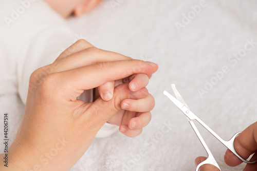 Closeup photo of mother's hands holding newborn's fingers and baby nail scissors on isolated white textile background
