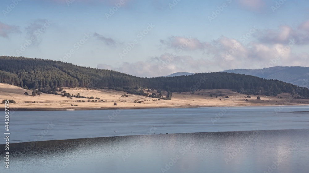 Natural landscape. Beautiful white clouds on blue sky and blurry reflections on the frozen water surface of Batak Dam, lake in the Rhodope Mountains, Bulgaria.