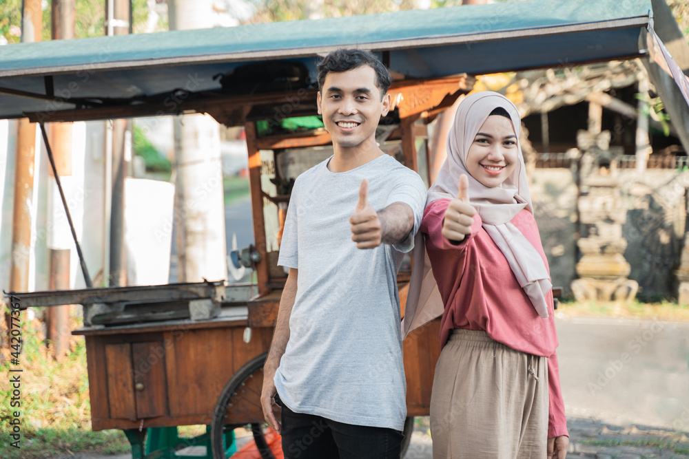 proud couple while selling food on the street. young muslim small business entrepreneur with their food cart at the background showing thumb up