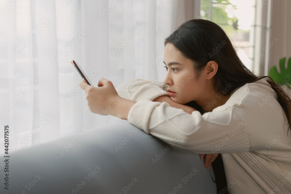 Young unhappy Asian woman using smart phone sitting on the couch with sadly face staying quarantine at her apartment. Lonely women sitting on a sofa looking out of the window. Young unhappy Asian woman using smart phone sitting on the couch with sadly face staying quarantine at her apartment. Lonely women sitting on a sofa looking out of the window.