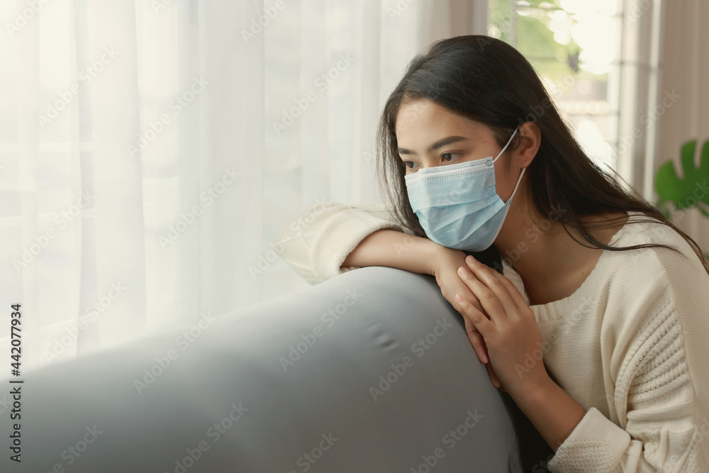 Young Asian woman wearing protective face mask stay quarantine at her apartment. Lonely women sitting on a couch looking out of the window