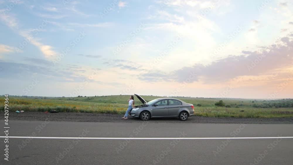 A frustrated young beautiful girl is standing by a broken car on the highway at sunset and calls the car service. Problems with the car. The girl waves her hands looking for help on the highway A frustrated young beautiful girl is standing by a broken car on the highway at sunset and calls the car service. Problems with the car. The girl waves her hands looking for help on the highway