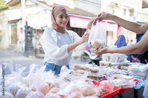 A beautiful girl in a veil buys takjil food in a plastic bag from a roadside stall seller