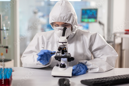 Scientist in ppe suit making adjustments and looking through laboratory microscope. Chemist in coverall working with various bacteria, tissue blood samples for antibiotics research.