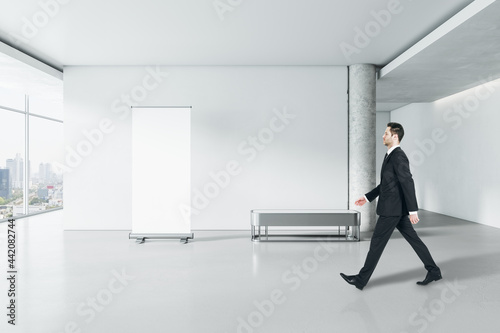 Businessman walking in modern gallery interior with empty poster on concrete wall and window with city view. Mock up.