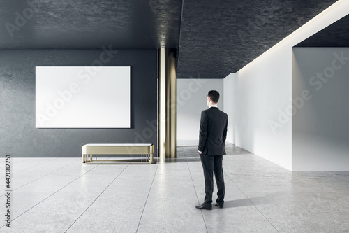 Businessman in modern gallery interior with empty poster on concrete wall. Mock up.