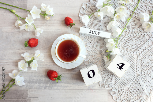 Calendar for July 4 : cubes with the numbers 0 and 4, the name of the month of July in English, a cup of tea on a gray openwork napkin, scattered flowers of bluebells and strawberries, top view