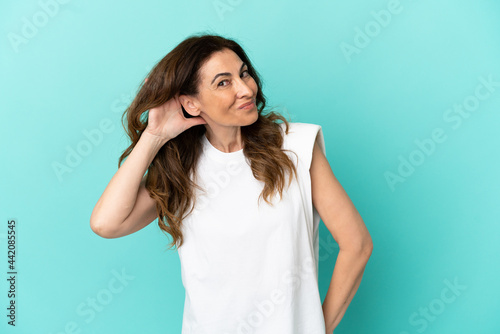 Middle aged caucasian woman isolated on blue background listening to something by putting hand on the ear