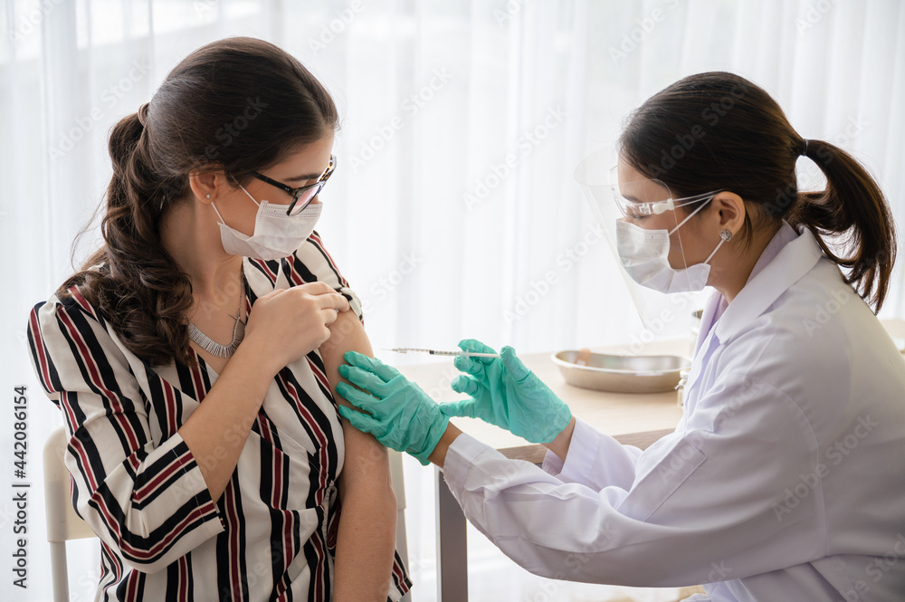 Female doctor or nurse wearing a mask, glove, and face shield are injecting the coronavirus 19 vaccine on shoulder of the woman to immunize in laboratory. Concept of preventing the spread of COVID-19. Female doctor or nurse wearing a mask, glove, and face shield are injecting the coronavirus 19 vaccine on shoulder of the woman to immunize in laboratory. Concept of preventing the spread of COVID-19.