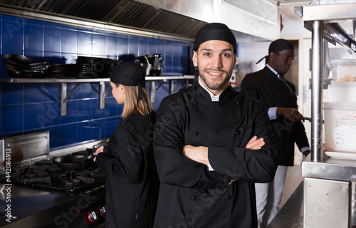 Confident chef of restaurant posing with arms crossed in kitchen on background with employees.